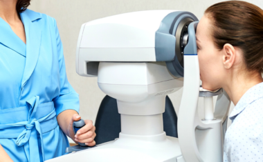 Image is of an optometrist standing before a medical device as a patient looks through the device and has her eyes checked.