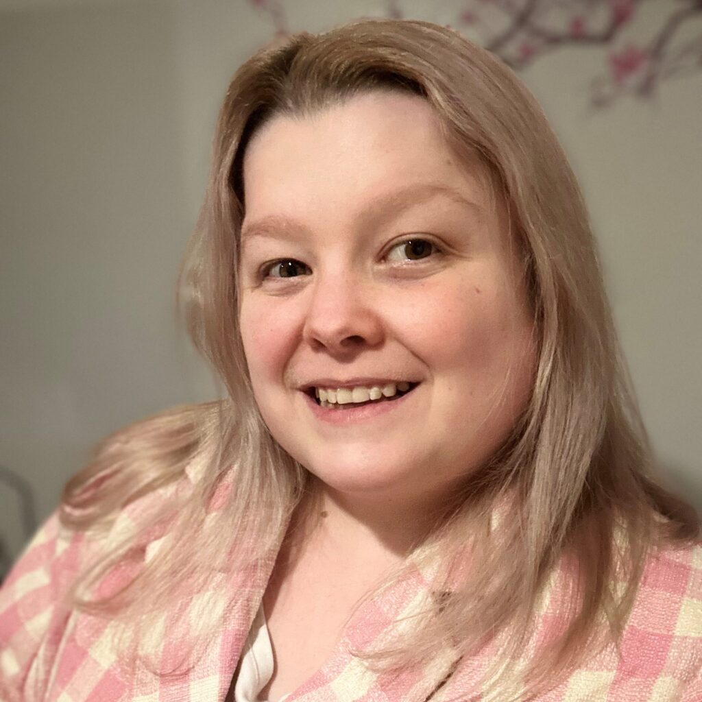 Photo of Alicia Chenier. Alicia is wearing a pink tartan blazer and is smiling at the camera.