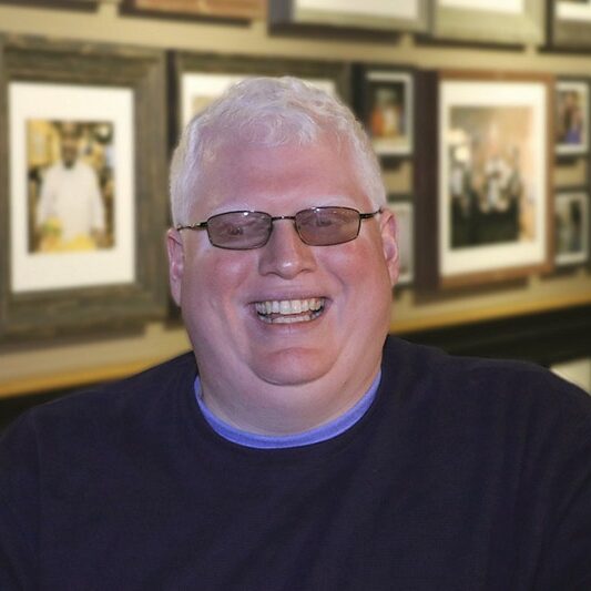 Photo of Dave Brown. Dave is in front of a wall of framed photographs. He is in a black sweater and is smiling at the camera.