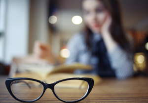 A pair of glasses on a table and a woman in the background reading a book